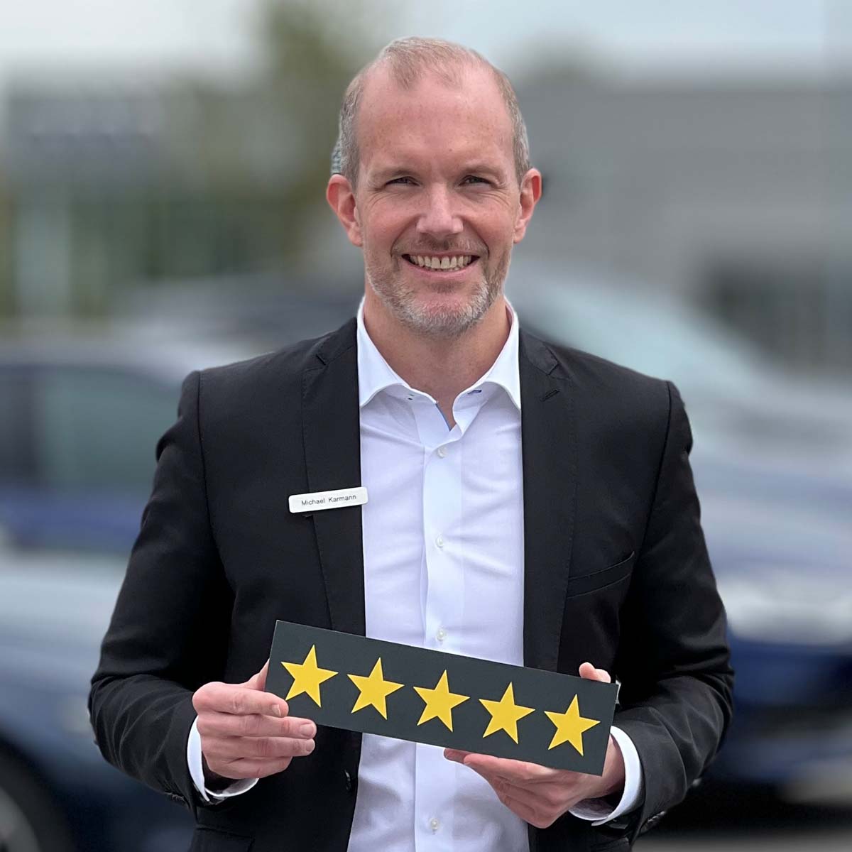 Gebrauchtwagenverkäufer Michael Karmann lächelt in die Kamera und hält ein Schild mit fünf Sternen in der Hand