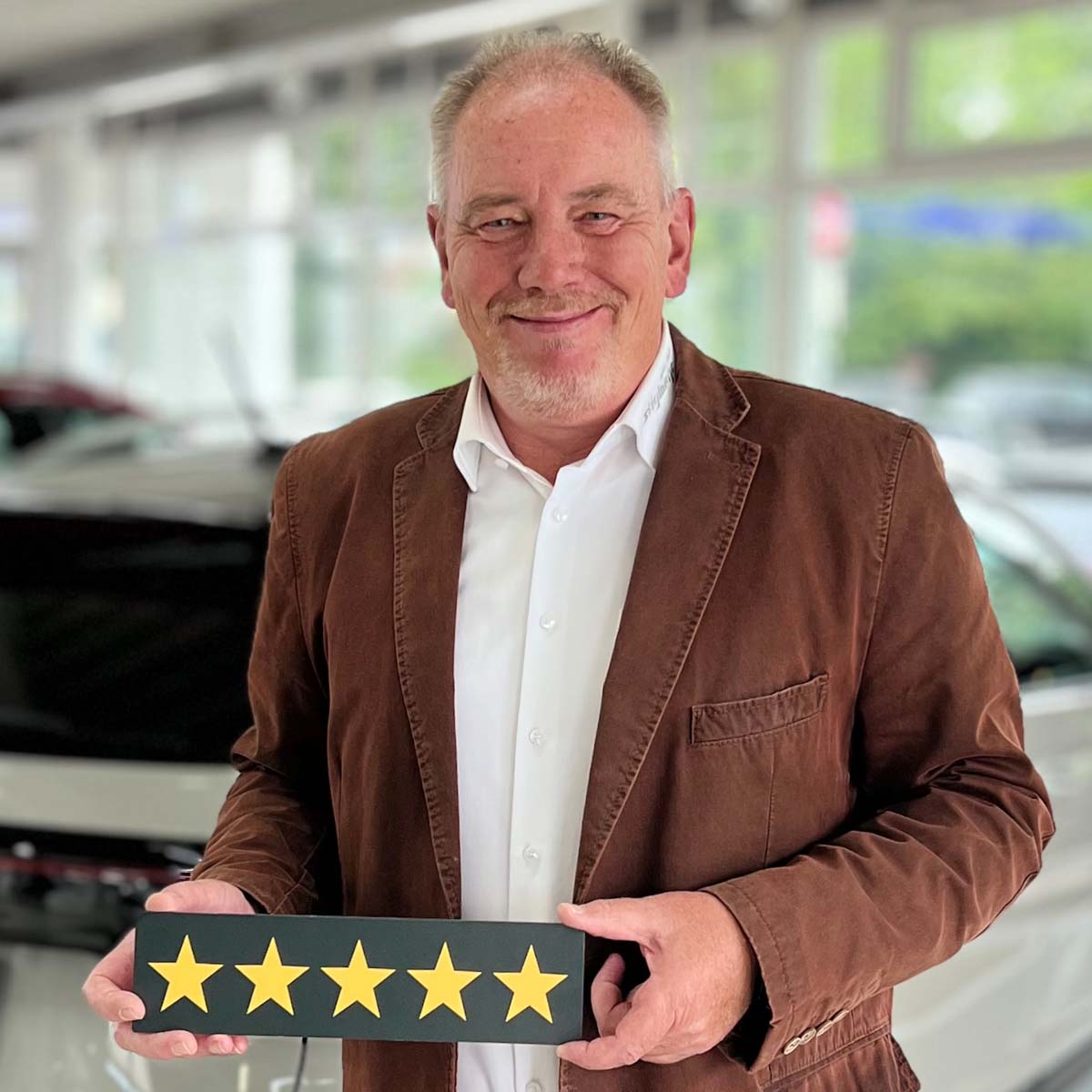 Neuwagen Verkäufer Norbert Pröhl lächelt in die Kamera und hält ein Schild mit fünf Sternen in der Hand